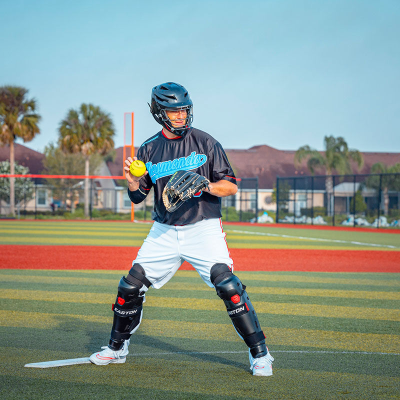 A pitcher on the mound wearing a black Easton Hellcat fielding helmet and leg guards - SKU: EHCATH